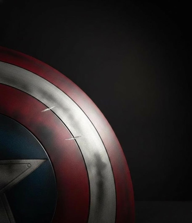 May include: A close-up of a Captain America shield. The shield features a red and silver striped design with a blue star emblem. The shield has a distressed, battle-worn appearance, with visible scratches and wear.