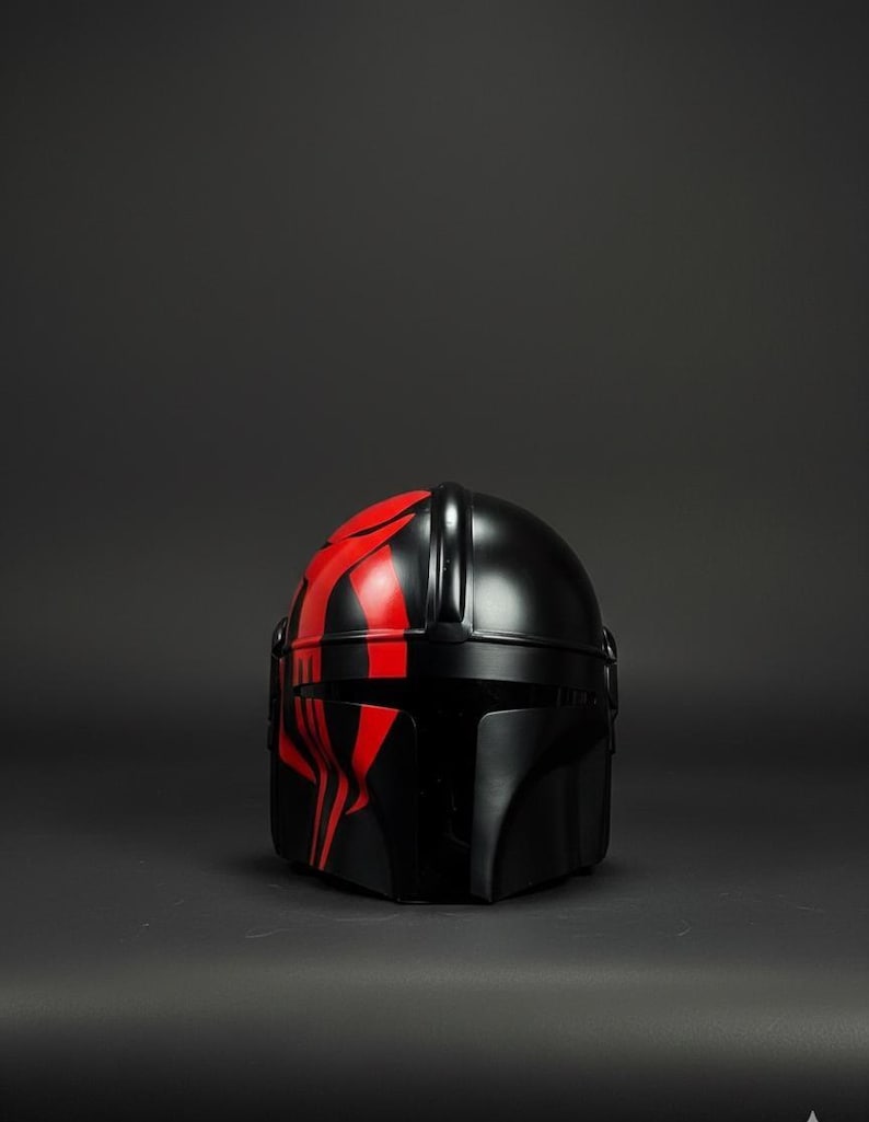 Op de afbeelding: Een zwarte helm met rode tribale markeringen op de voorkant en bovenkant. De helm heeft een gladde, glanzende afwerking en een afgeronde vorm, met een vizier en andere details. Het is waarschijnlijk een kostuumstuk of verzamelobject.