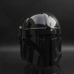 May include: A shiny black helmet, similar to a Mandalorian design, is shown against a dark backdrop. The helmet has a rounded crown, a visor, and angular cheek plates. It rests on a wireframe stand.