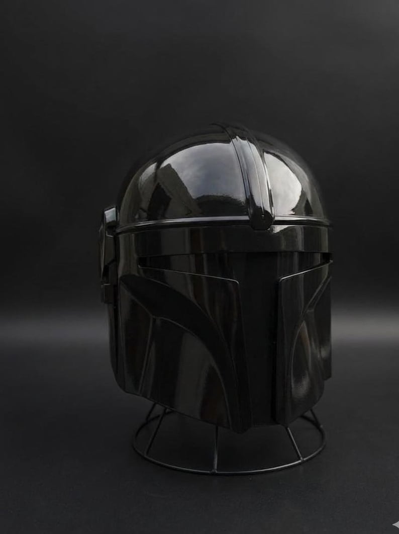 May include: A shiny black helmet, similar to a Mandalorian design, is shown against a dark backdrop. The helmet has a rounded crown, a visor, and angular cheek plates. It rests on a wireframe stand.