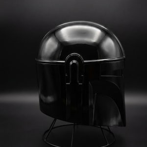 May include: A shiny black helmet, possibly a prop or collectible, is shown against a dark backdrop. The helmet has a rounded crown and angular visor, with a distinctive T-shaped visor and a wire stand.