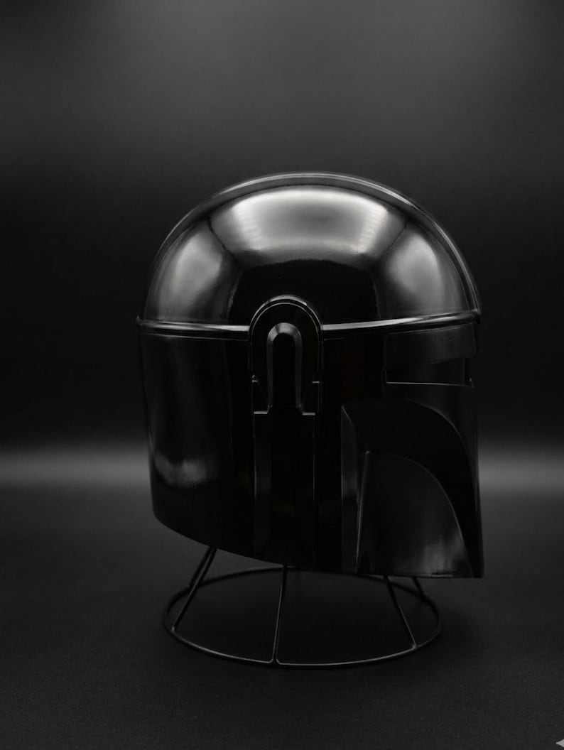 May include: A shiny black helmet, possibly a prop or collectible, is shown against a dark backdrop. The helmet has a rounded crown and angular visor, with a distinctive T-shaped visor and a wire stand.