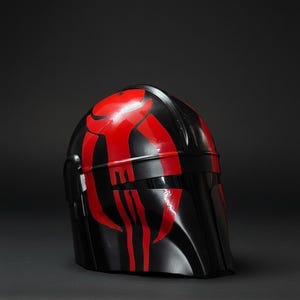 Op de afbeelding: Een zwarte helm met rode accenten. De helm heeft een glanzende afwerking en een opvallend ontwerp. Het rode ontwerp is symmetrisch en gedurfd, contrasterend met de zwarte basis. Waarschijnlijk een kostuumstuk of verzamelobject.