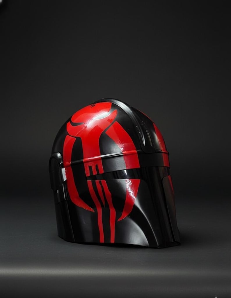 Op de afbeelding: Een zwarte helm met rode accenten. De helm heeft een glanzende afwerking en een opvallend ontwerp. Het rode ontwerp is symmetrisch en gedurfd, contrasterend met de zwarte basis. Waarschijnlijk een kostuumstuk of verzamelobject.