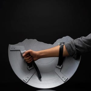 Puede incluir: Un escudo semicircular gris con un asa y una correa para el brazo. El escudo tiene una superficie texturizada y le falta una secci&oacute;n en la parte superior. El escudo lo sostiene una persona.