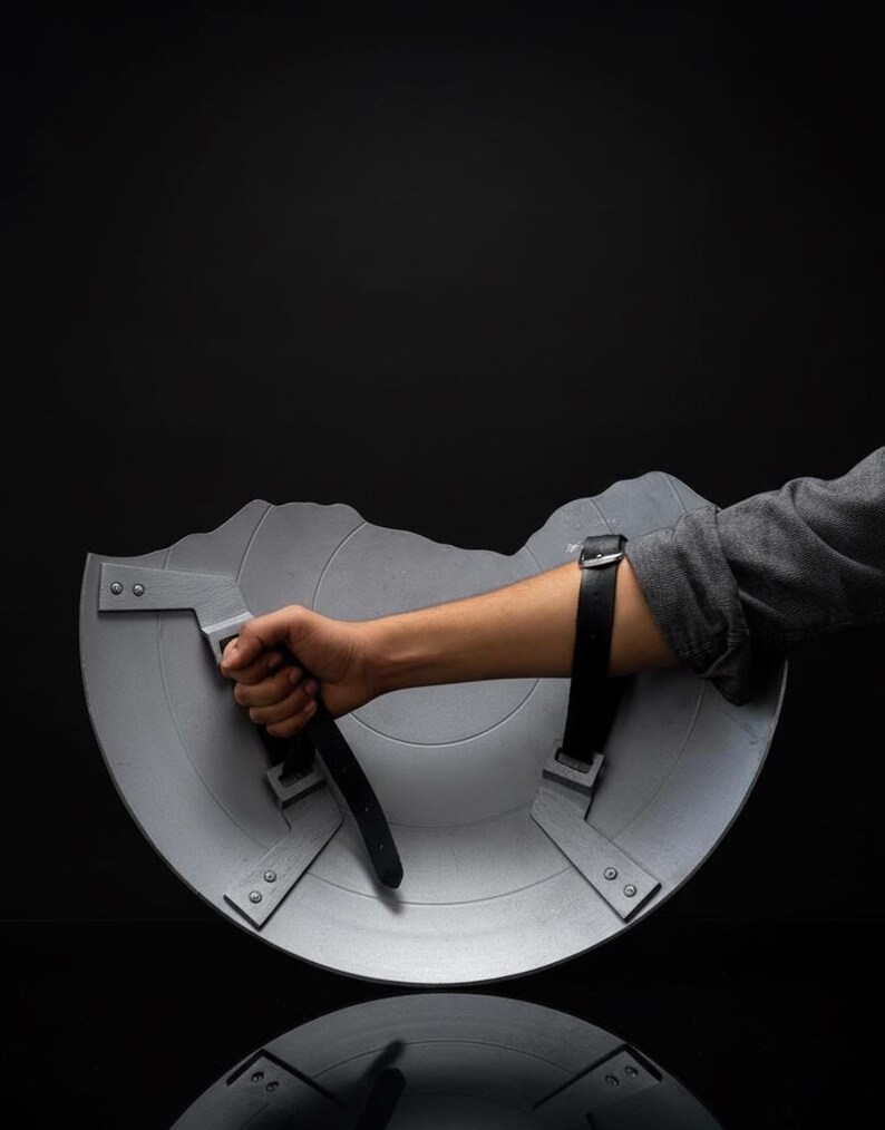Puede incluir: Un escudo semicircular gris con un asa y una correa para el brazo. El escudo tiene una superficie texturizada y le falta una secci&oacute;n en la parte superior. El escudo lo sostiene una persona.