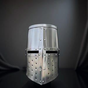 Puede incluir: Un yelmo de caballero medieval plateado. El casco tiene una parte superior plana, aberturas rectangulares para los ojos y numerosos agujeros de ventilación. Remaches aseguran las placas de metal. Se muestra sobre un soporte negro.