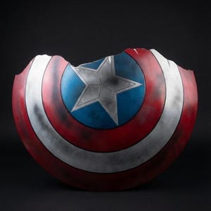 Puede incluir: Un escudo de Capit&aacute;n Am&eacute;rica da&ntilde;ado, con una combinaci&oacute;n de colores rojo, blanco y azul. El escudo tiene una estrella en el centro y un borde roto, lo que le da una apariencia desgastada por la batalla. Un objeto de colecci&oacute;n o utiler&iacute;a.