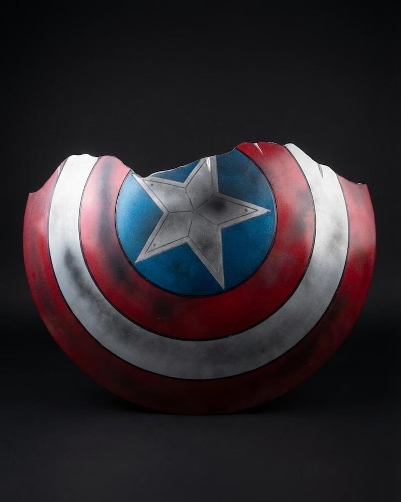 Puede incluir: Un escudo de Capit&aacute;n Am&eacute;rica da&ntilde;ado, con una combinaci&oacute;n de colores rojo, blanco y azul. El escudo tiene una estrella en el centro y un borde roto, lo que le da una apariencia desgastada por la batalla. Un objeto de colecci&oacute;n o utiler&iacute;a.