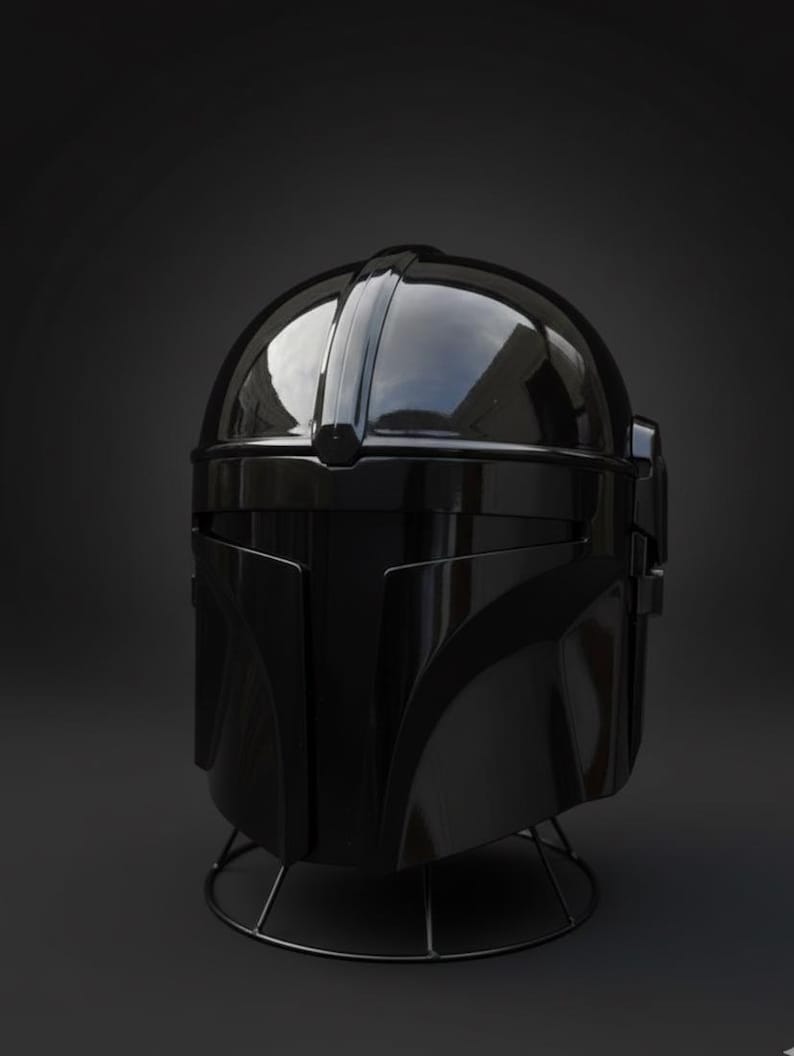 May include: A shiny black helmet with a rounded crown and sharp angles. The helmet features a visor and cheek plates, and it rests on a black wire stand. The design hints at a futuristic or science fiction aesthetic.