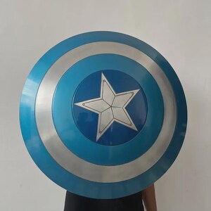 May include: A circular shield featuring a blue and silver design. The shield has a large white star at its centre, encircled by bands of blue and silver. This shield is likely a prop or costume accessory.