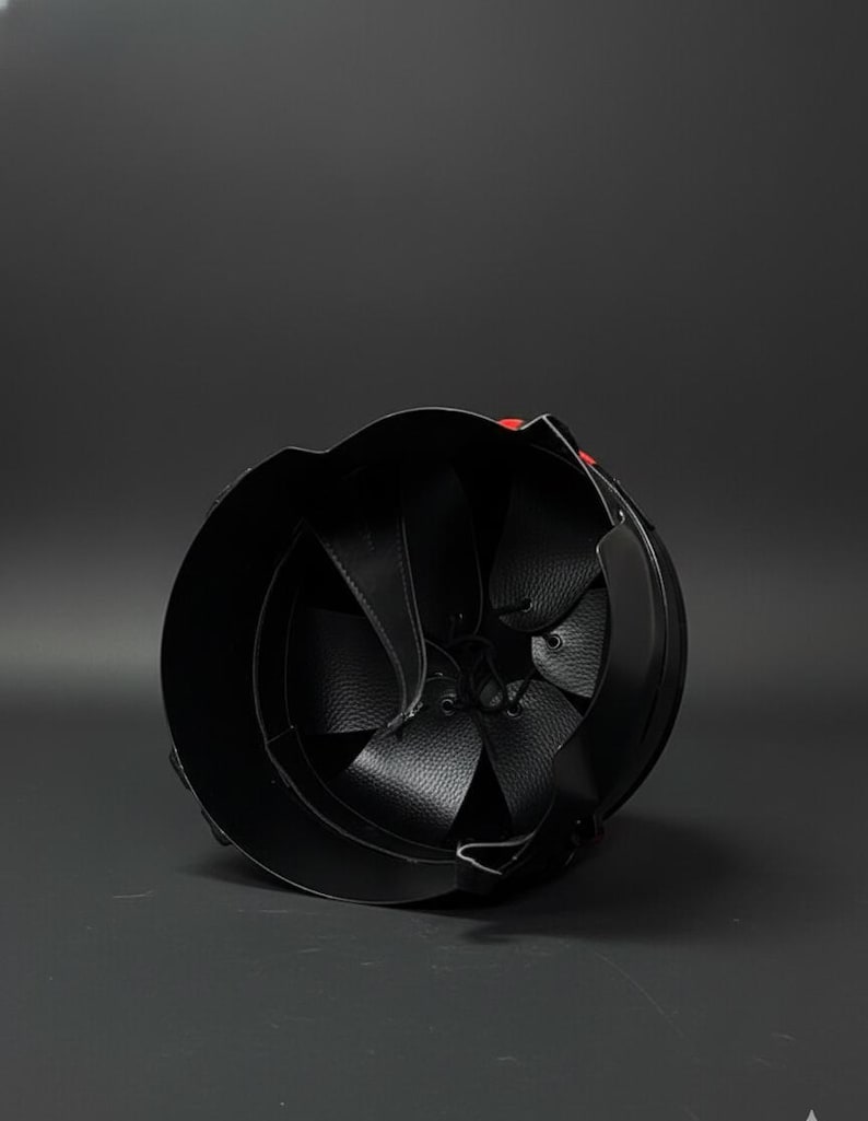 Op de afbeelding: Zwarte leren helm met een afgeronde vorm en een gedetailleerd interieur. Het interieur heeft leren panelen en riemen. Een klein rood accent is zichtbaar aan de buitenkant. De helm is ontworpen voor bescherming.
