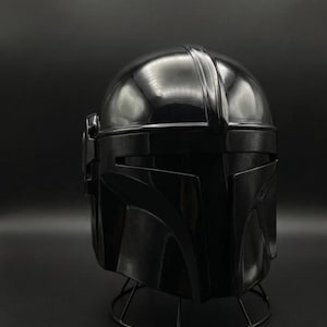 May include: A black Mandalorian helmet, a replica of the iconic headgear from the Star Wars series. The helmet is glossy and sits on a wireframe stand, showcasing its detailed design and shape.