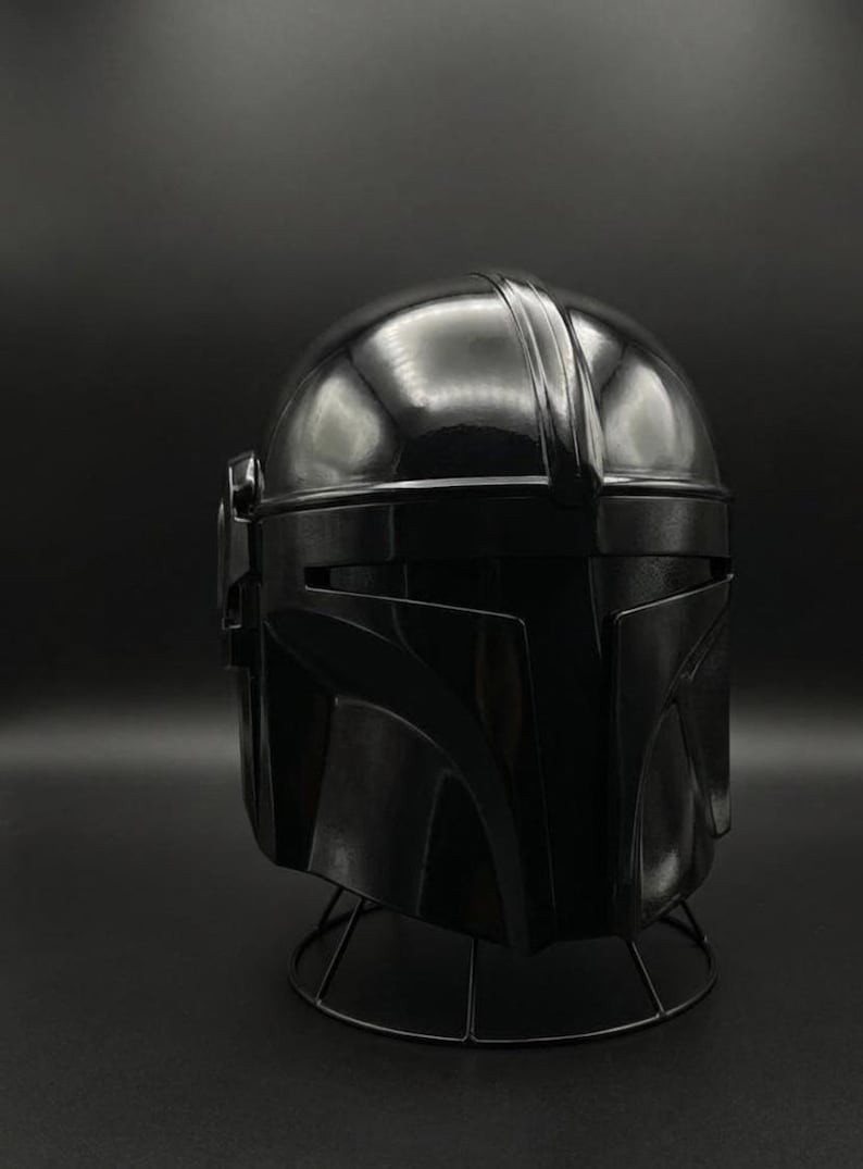 May include: A black Mandalorian helmet, a replica of the iconic headgear from the Star Wars series. The helmet is glossy and sits on a wireframe stand, showcasing its detailed design and shape.
