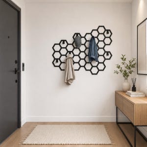 May include: A black metal wall-mounted coat rack featuring a geometric honeycomb pattern. The rack includes multiple integrated hooks for hanging items, with a grey baseball cap, a beige jacket, and a blue denim jacket currently hanging on it.