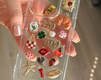 Puede incluir: Funda transparente para teléfono con abalorios dorados y coloridos. Los abalorios incluyen un anillo dorado, un corazón, un cruasán, un trébol de cuatro hojas, una fresa, una cereza, una estrella de mar y las letras "JSW" y "BTW".