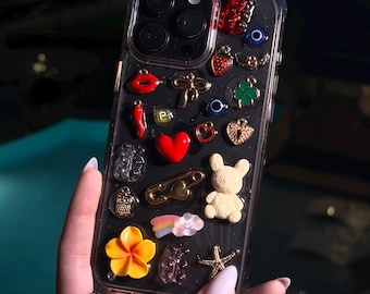 Puede incluir: Una funda transparente para teléfono adornada con varios dijes, incluyendo un corazón rojo, un imperdible dorado, una flor amarilla y un oso de peluche blanco. Otros dijes incluyen un chile rojo, una fresa y un trébol de cuatro hojas.