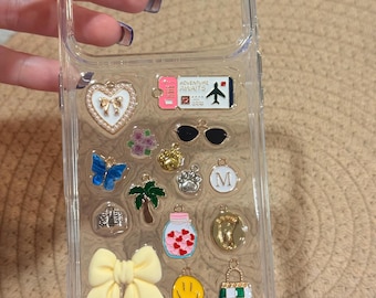 Puede incluir: Una funda de teléfono transparente decorada con varios dijes pequeños, incluyendo un corazón con un lazo, un billete de avión rosa con la etiqueta Adventure Awaits, gafas de sol, una mariposa azul, una palmera, un frasco de corazones, un lazo amarillo, una cara sonriente y un bolso.