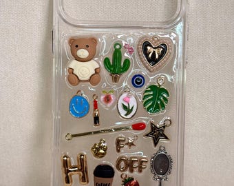 Puede incluir: Funda de teléfono transparente decorada con varios dijes, incluyendo un oso de peluche, un cactus, un corazón, una carita sonriente, un lápiz labial y una taza de café. Otros dijes incluyen una fresa, una estrella y un pequeño espejo. La funda tiene forma rectangular.
