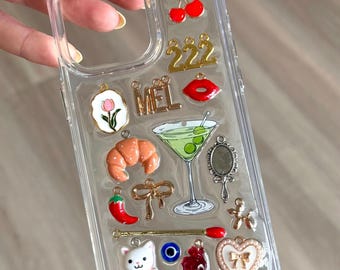 Puede incluir: Funda de teléfono transparente decorada con varios dijes, incluyendo cerezas, un cruasán, un martini y un gato. Otros dijes incluyen un chile rojo, un corazón y el número 222. La funda tiene una variedad de colores y texturas.