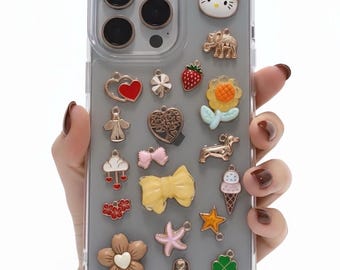 Puede incluir: Una funda transparente para teléfono adornada con varios dijes. Los dijes incluyen un Hello Kitty, un elefante, un corazón, una fresa, una flor, un perro salchicha, un helado, un lazo y un trébol. La funda está sujeta por una mano.