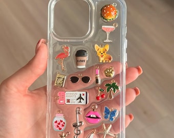 Puede incluir: Funda transparente para teléfono adornada con varios dijes, incluyendo una hamburguesa, cóctel, flamenco, taza de café, gafas de sol, lápiz labial y un billete que dice "Adventure Awaits". Otros dijes incluyen un perro, cerezas, una palmera, una mariposa y una estrella de mar.