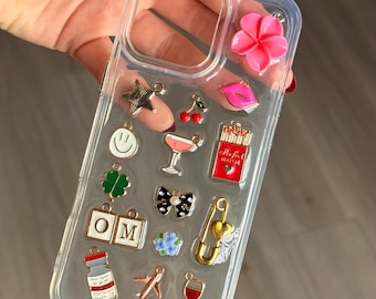 Puede incluir: Funda transparente para teléfono adornada con varios dijes, que incluyen una flor rosa, una estrella, cerezas, labios, una carita sonriente, un trébol de cuatro hojas, un lazo, las letras "OM", un vial, un avión, un cóctel, una caja de cerillas, un imperdible y una copa de vino.