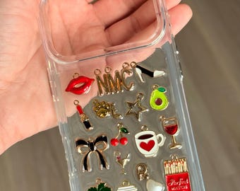 Puede incluir: Funda transparente para teléfono con varios dijes, incluyendo un labial rojo, un lazo negro, una taza de café y una caja de cerillas roja. Otros dijes incluyen un tacón alto, aguacate y una copa de vino.