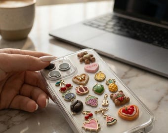 Puede incluir: Funda transparente para teléfono decorada con varios dijes, incluyendo un oso de peluche, cerezas, un corazón con el texto "xoxo" y una rodaja de sandía. Los dijes son de varios colores y formas.