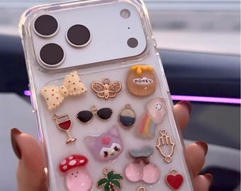 Puede incluir: Funda de teléfono transparente decorada con varios dijes, incluyendo un lazo, una mariposa, gafas de sol, un tarro de miel, un arcoíris, un gato, una seta, una flor, una corona, un osito de peluche y un corazón.