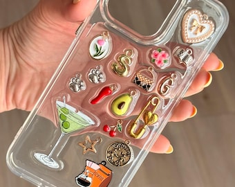 Puede incluir: Funda transparente para teléfono con varios dijes, incluyendo una copa de martini, una taza de café, un corazón y un imperdible dorado. Los dijes son de varios colores y formas, agregando un toque decorativo a la funda.