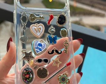 Puede incluir: Funda transparente para teléfono decorada con varios dijes, incluyendo una llave, un corazón, una estrella y una mariposa. Los dijes son plateados, dorados, rojos y azules. La funda se sostiene contra un fondo borroso al aire libre.