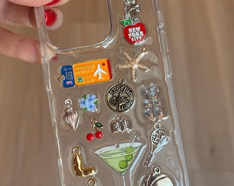 Puede incluir: Funda de teléfono transparente decorada con varios dijes, incluyendo una Estatua de la Libertad con una manzana, una estrella de mar, una copa de martini y una pelota de baloncesto. Otros dijes incluyen un pasaporte, flores, cerezas y las letras "J" y "L".