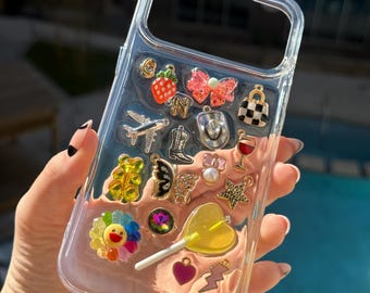 Puede incluir: Funda transparente para teléfono decorada con varios abalorios, incluyendo una fresa, un lazo, un avión, un sombrero de vaquero, una copa de vino y un girasol. La funda también presenta una piruleta amarilla en forma de corazón y un charm de corazón rosa.