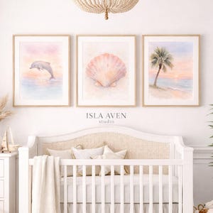 Puede incluir: Tres impresiones en acuarela enmarcadas con temas de playa. Una muestra un delfín saltando del agua, otra una concha y la tercera una palmera en una playa. Las impresiones están sobre una cuna blanca. El texto "ISLA AVEN studio" está debajo.
