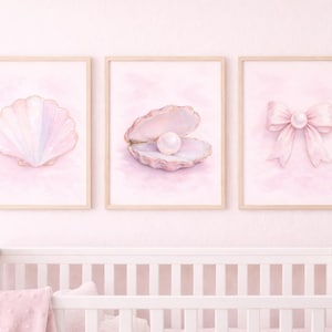 Op de afbeelding: Drie ingelijste aquarelprints met een babymotief. De prints tonen een zeeschelp, een openstaande schelp met een parel en een strik met een parel. Het kleurenpalet is zachtroze en goud, perfect voor een kinderkamer.