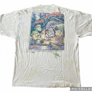 Puede incluir: Camiseta de color crema con un gr&aacute;fico colorido de animales de dibujos animados acampando. La imagen incluye una tortuga tocando la guitarra, osos alrededor de una fogata y el texto "LIKE RAIN". Tambi&eacute;n se ve una rana.
