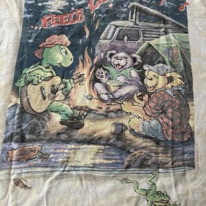 Puede incluir: Camiseta blanquecina con una escena de acampada con personajes de dibujos animados. Una tortuga toca la guitarra junto a una fogata con osos y una furgoneta al fondo. El texto dice "...Feels Like Rain". Tambi&eacute;n se ven una rana y un pez.