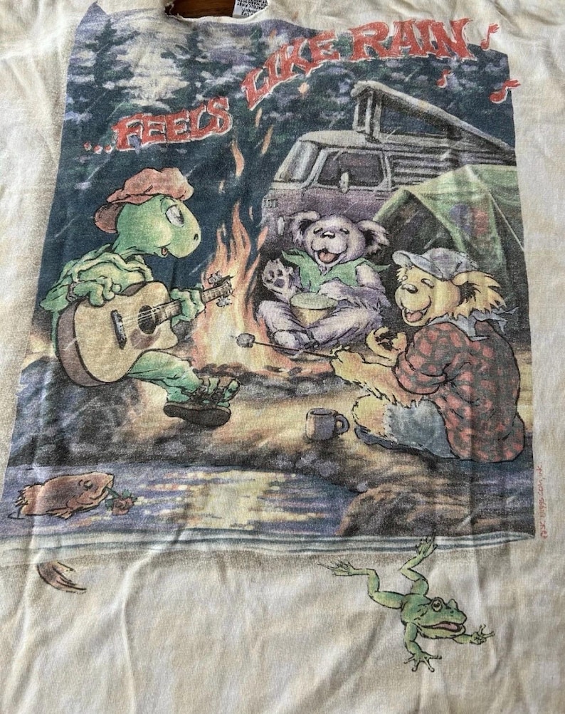 Puede incluir: Camiseta blanquecina con una escena de acampada con personajes de dibujos animados. Una tortuga toca la guitarra junto a una fogata con osos y una furgoneta al fondo. El texto dice "...Feels Like Rain". Tambi&eacute;n se ven una rana y un pez.