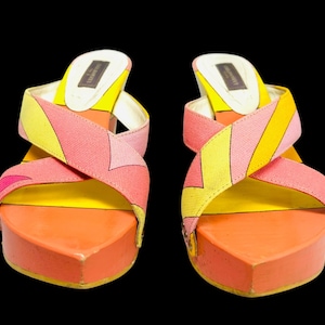 May include: A pair of colorful platform sandals with a geometric pattern in pink, yellow, and orange. The sandals have a thick, orange platform sole and criss-cross straps. The background is black.