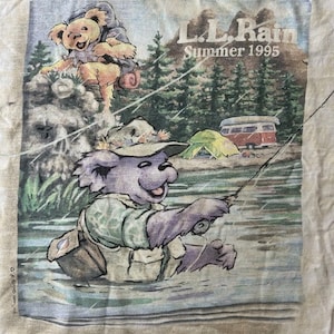 Puede incluir: Camiseta blanquecina con un gr&aacute;fico de un oso pescando en un r&iacute;o, con el texto "L.L. Rain Summer 1995". El oso lleva un sombrero y un chaleco de pesca. Una tienda de campa&ntilde;a y una furgoneta camper est&aacute;n en el fondo.