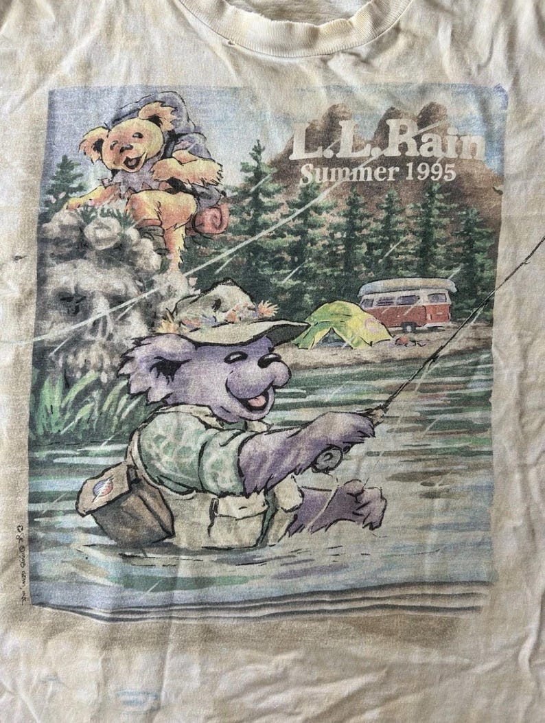 Puede incluir: Camiseta blanquecina con un gr&aacute;fico de un oso pescando en un r&iacute;o, con el texto "L.L. Rain Summer 1995". El oso lleva un sombrero y un chaleco de pesca. Una tienda de campa&ntilde;a y una furgoneta camper est&aacute;n en el fondo.