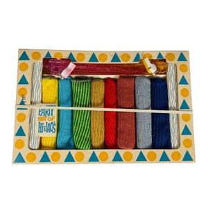 May include: A wooden box containing a rainbow set of "Hi-Jacs" fabric strips. The box has a yellow background with blue and orange triangles. The fabric strips are in a variety of colours, including red, orange, yellow, green, blue, and purple.