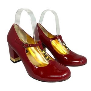 May include: Pair of red patent leather Mary Jane heels with gold-tone accents. The shoes feature a T-strap design with a buckle closure and a block heel with a gold-tone detail. The interior is gold-colored.