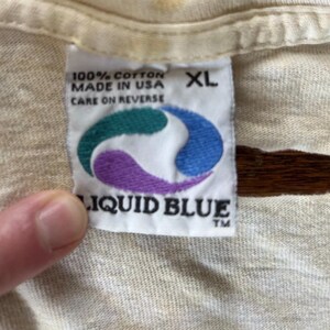 Puede incluir: Primer plano de una etiqueta de camiseta color crema. La etiqueta dice "100% Cotton Made in USA Care on Reverse" y presenta un logotipo turquesa, azul y morado con las palabras "Liquid Blue" debajo. La etiqueta tambi&eacute;n indica la talla XL.