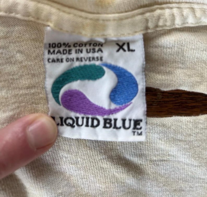 Puede incluir: Primer plano de una etiqueta de camiseta color crema. La etiqueta dice "100% Cotton Made in USA Care on Reverse" y presenta un logotipo turquesa, azul y morado con las palabras "Liquid Blue" debajo. La etiqueta tambi&eacute;n indica la talla XL.