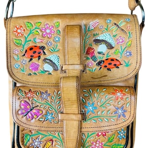 May include: Tan leather crossbody bag with hand-painted floral, mushroom, ladybug, and butterfly designs. The bag has two front pockets and a flap closure with a buckle. The strap is adjustable.
