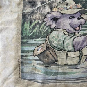 Puede incluir: Una camiseta de color claro con un oso de dibujos animados pescando en un r&iacute;o. El oso lleva un sombrero, un chaleco y una bandolera. La imagen tiene un estilo de acuarela con tonos verdes, azules y marrones. El texto "&copy; & &copy; 1995 Grateful Dead Merch, Inc." est&aacute; impreso en el lado izquierdo.