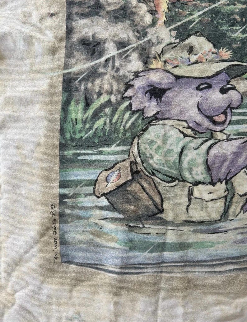 Puede incluir: Una camiseta de color claro con un oso de dibujos animados pescando en un r&iacute;o. El oso lleva un sombrero, un chaleco y una bandolera. La imagen tiene un estilo de acuarela con tonos verdes, azules y marrones. El texto "&copy; & &copy; 1995 Grateful Dead Merch, Inc." est&aacute; impreso en el lado izquierdo.