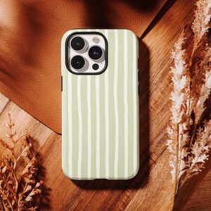 May include: A light green phone case with a vertical white stripe pattern. The case has a black border and a camera cutout. The case is on a wooden surface with dried plants and a leather background.